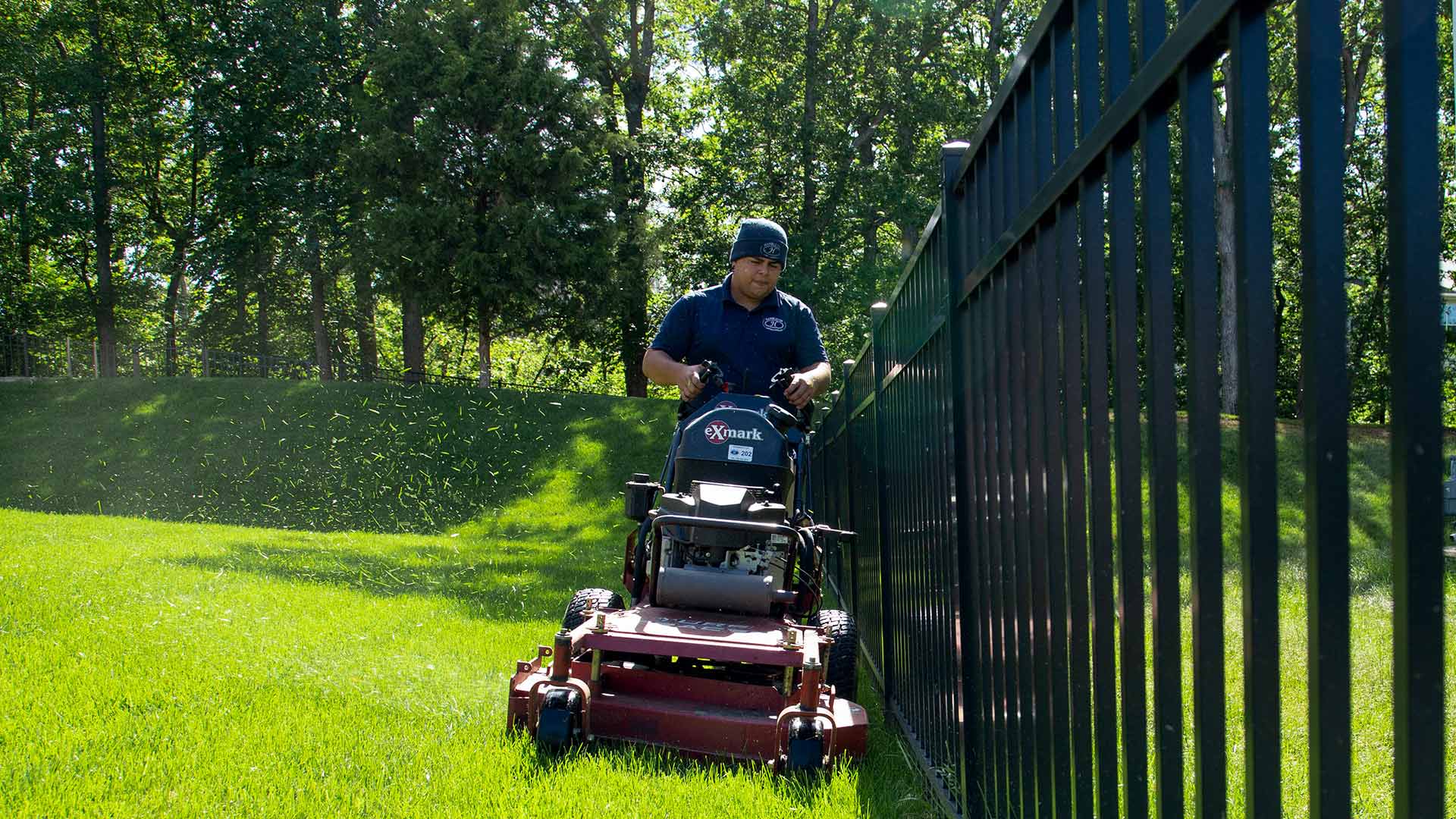 A worker is mowing a lawn by a fence with a line of trees in the background in Fairfax, VA.
