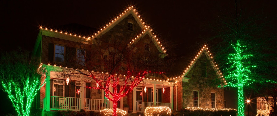 A house with holiday lighting along the windows, doorway, roofline, shrubs and small trees.