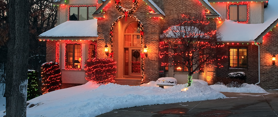A house with holiday lighting along the windows, doorway, roofline, shrubs and small trees.