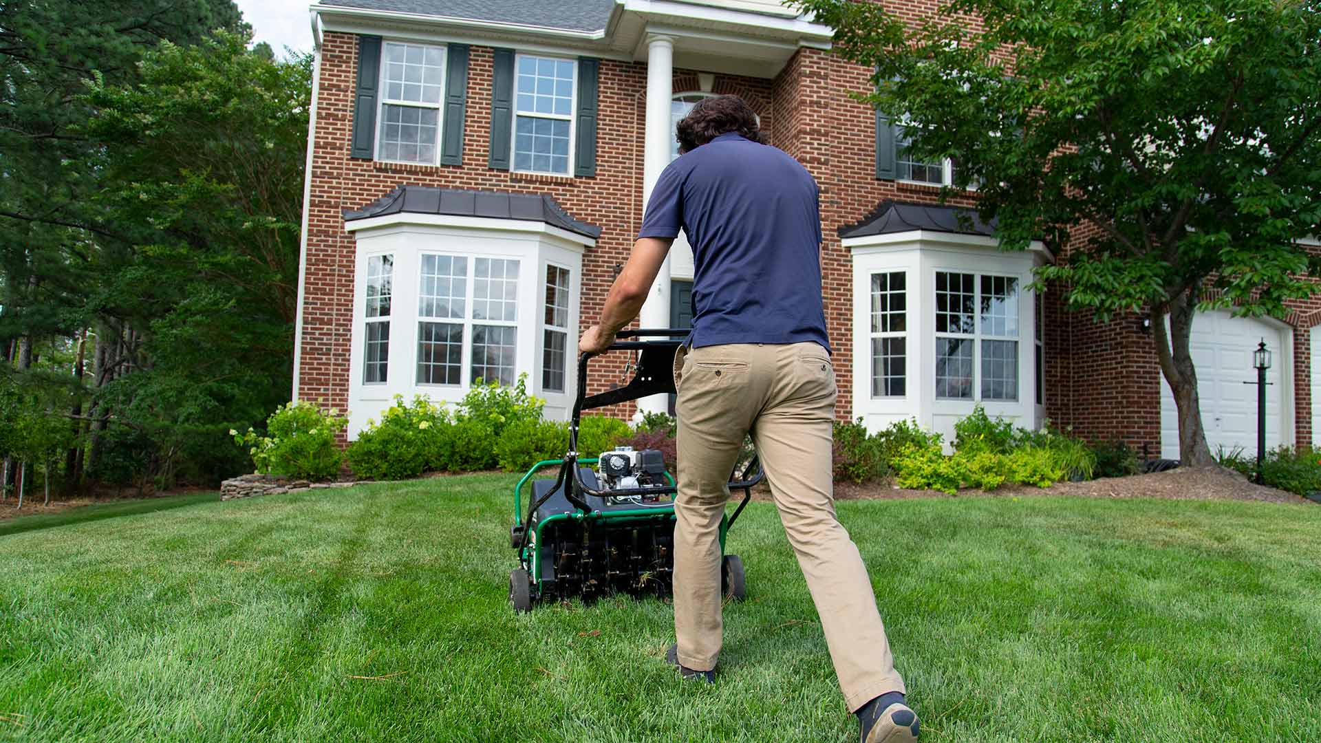 A man aerating a beautiful lawn with a brick house in the background.